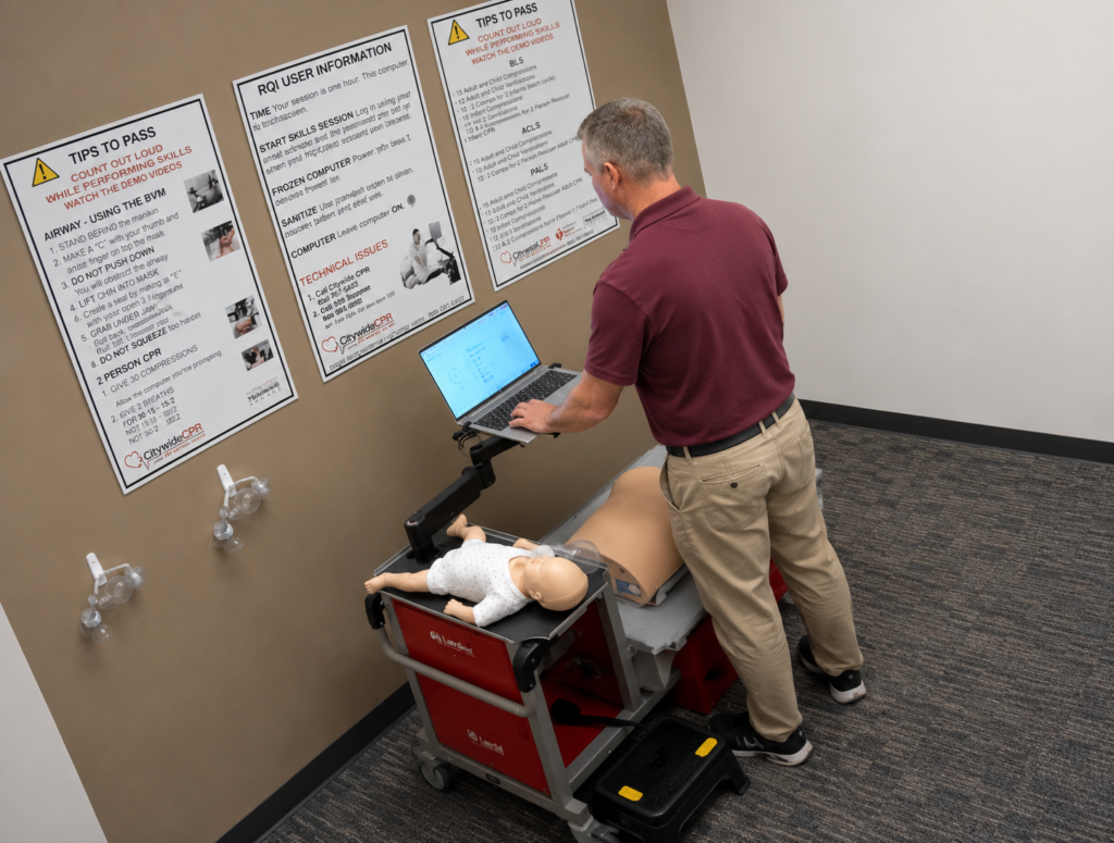 Participant using a laptop at the Citywide CPR simulation station during American Heart Association BLS skills training in Ann Arbor, Michigan, with adult and infant CPR manikins visible.