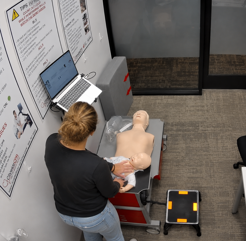 Participant performing infant CPR chest compressions on an infant CPR manikin at the Citywide CPR simulation station during American Heart Association BLS skills training in Arlington, Texas, with an adult CPR manikin and laptop visible.