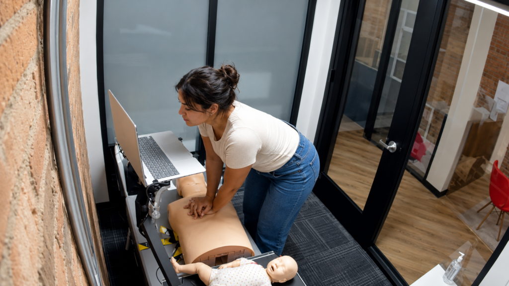 Participant performing chest compressions on an adult CPR manikin at a Citywide CPR simulation station in Baltimore, Maryland, with a laptop and infant manikin in a onesie visible.
