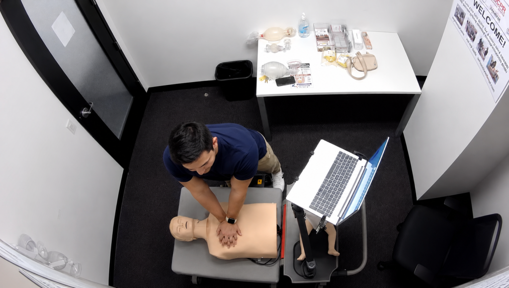 Participant performing chest compressions on an adult CPR manikin beside a laptop at the Citywide CPR simulation station in Beacon Hill, Boston.