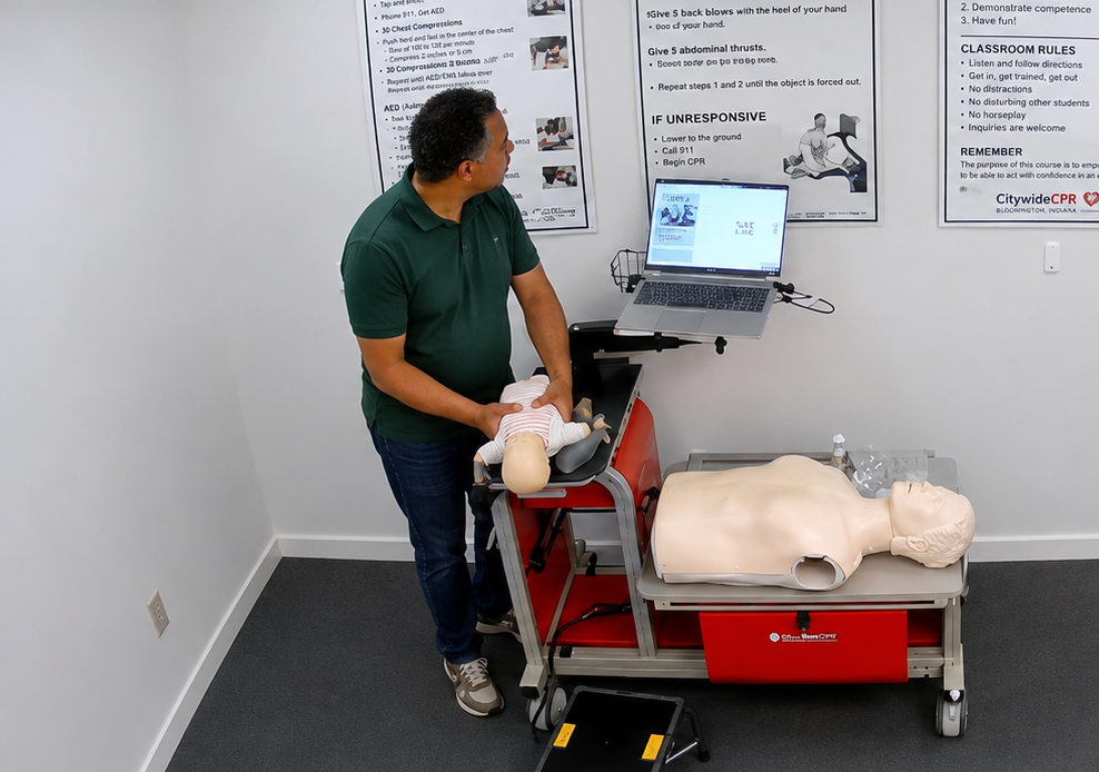 Participant holding an infant CPR manikin in a onesie at a Citywide CPR simulation station in Bloomington, Indiana, with an adult CPR manikin and laptop visible.