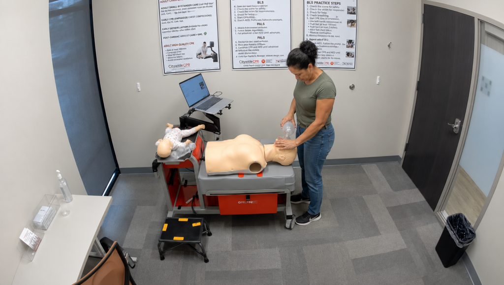 Participant providing bag-valve-mask ventilation to an adult CPR manikin at a Citywide CPR simulation station in Cedar Rapids, Iowa, with an infant manikin in a onesie and laptop visible.