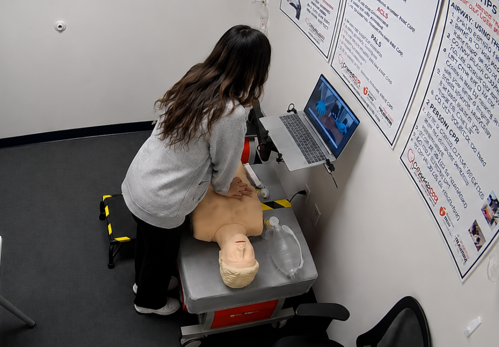 Participant performing chest compressions on an adult CPR manikin at a Citywide CPR training station in Dallas, Texas, with a laptop and CPR training equipment visible.