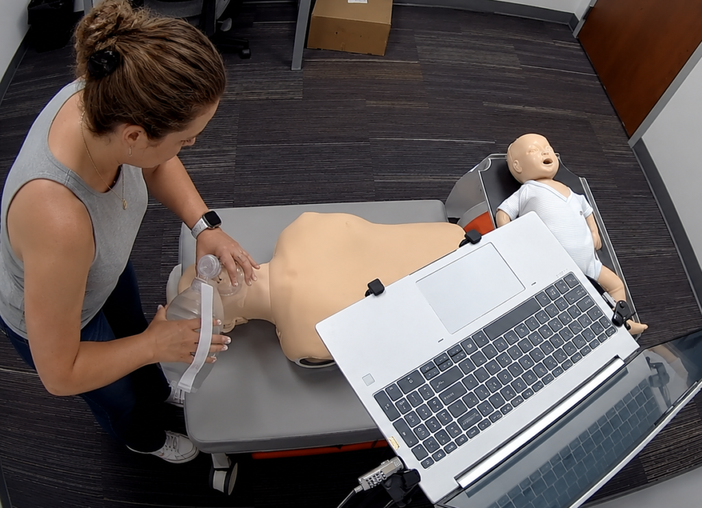 Participant providing bag-valve-mask ventilation to an adult CPR manikin at a Citywide CPR training station in Elgin, Illinois, with an infant CPR manikin wearing a white onesie and a laptop visible.
