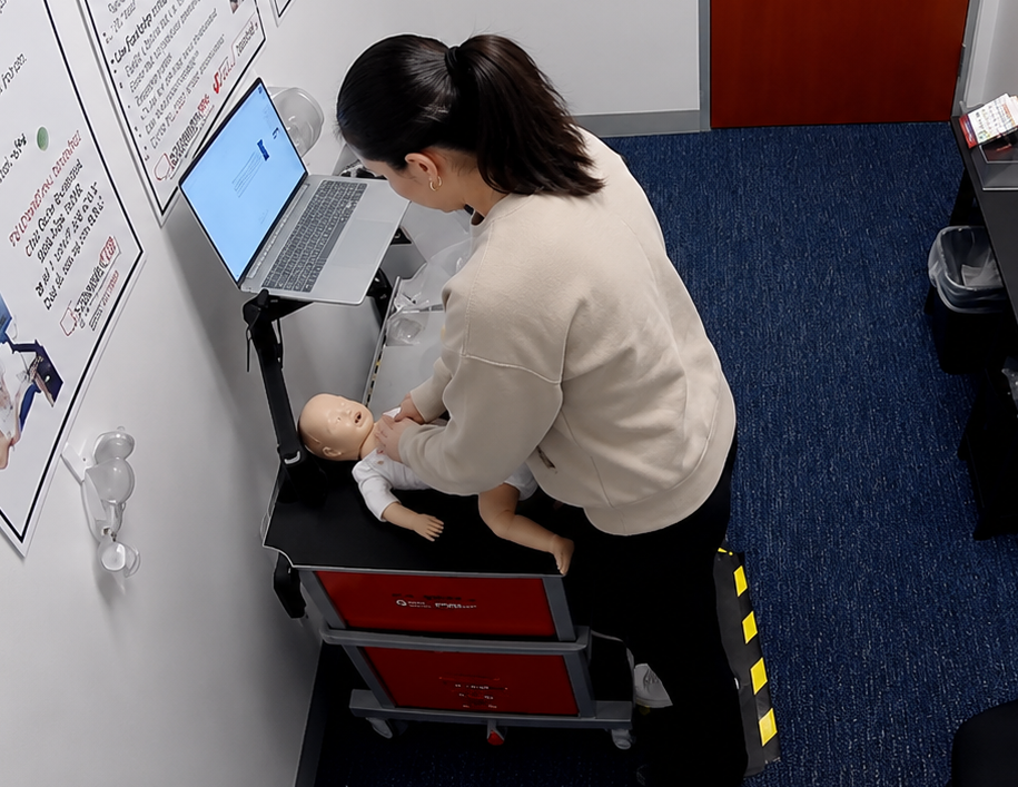 Participant practicing chest compressions on an infant CPR manikin beside a laptop and instructional posters at a Citywide CPR training station in Fort Worth, Texas.