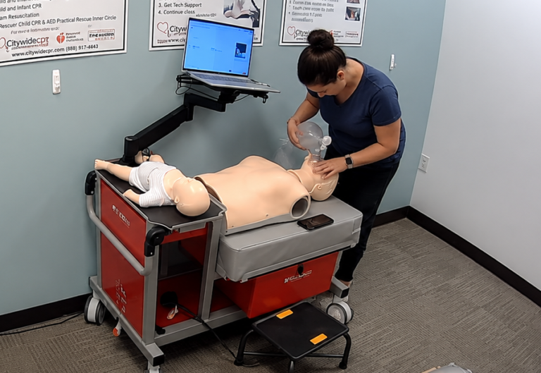 Participant providing bag-valve-mask ventilation to an adult CPR manikin at a Citywide CPR training station in Hartford, Connecticut, with an infant CPR manikin wearing a white onesie and a laptop visible.