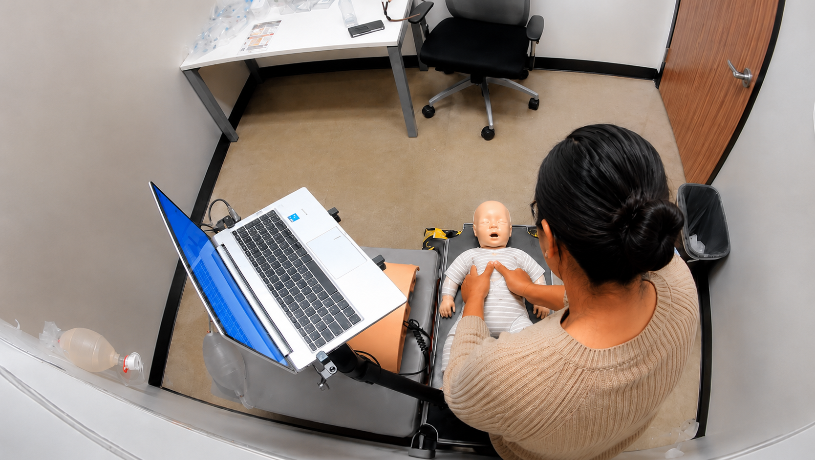 Participant performing chest compressions on a fully clothed infant CPR manikin beside a laptop at a Citywide CPR training station in Hyde Park, Chicago.