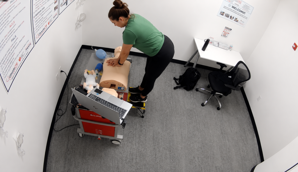 Participant performing chest compressions on an adult CPR manikin at a Citywide CPR training station in Jacksonville, Florida, with an infant CPR manikin wearing a white onesie, a laptop, and CPR training equipment visible.