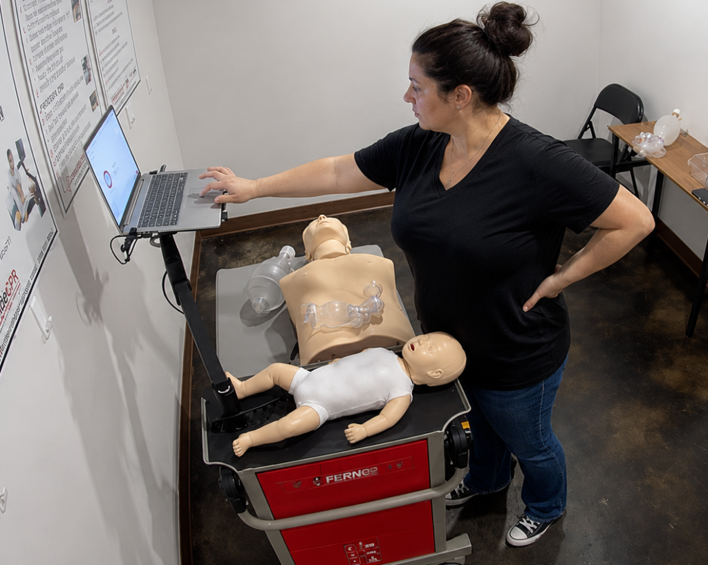 Participant using a laptop at a Citywide CPR training station in Lincoln, Nebraska, with an adult CPR manikin, an infant manikin in a white onesie, and airway equipment visible.