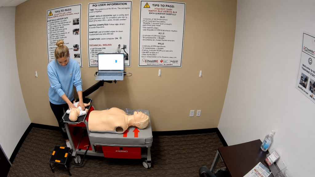 Participant performing chest compressions on an infant CPR manikin at a Citywide CPR training station in Louisville, Kentucky, with an adult CPR manikin and laptop visible.
