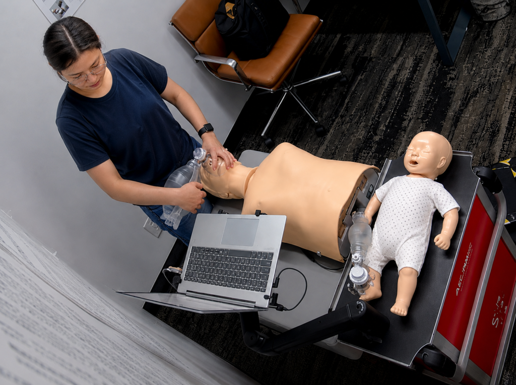 Participant providing bag-valve-mask ventilation to an adult CPR manikin at a Citywide CPR training station in Midtown Manhattan, New York, with an infant CPR manikin wearing a white onesie and a laptop visible.