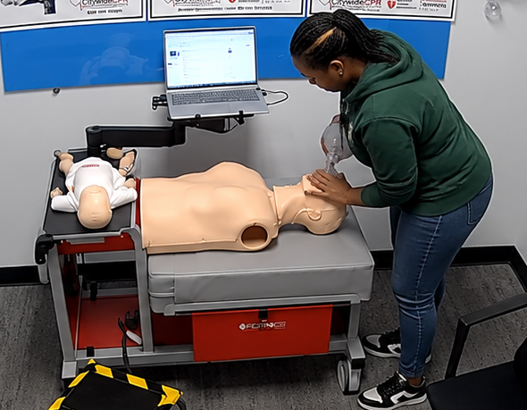 Participant providing bag-valve-mask ventilation to an adult CPR manikin at a Citywide CPR training station in Milwaukee, Wisconsin, with an infant manikin in a white onesie and a laptop visible.