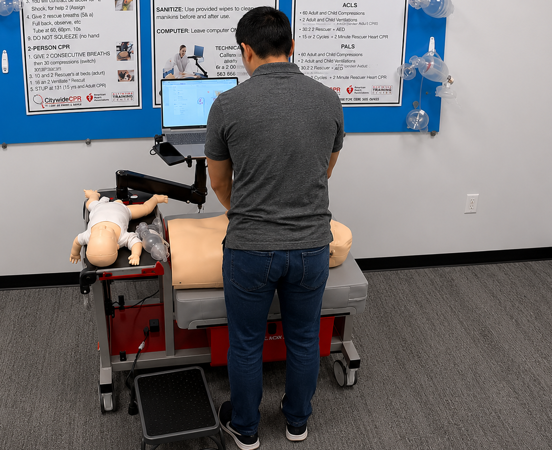 American Heart Association CPR Certification Training in Minneapolis, Minnesota Participant standing at a CPR skills station using a laptop while completing American Heart Association adult and infant CPR training with adult and infant manikins in Minneapolis, Minnesota.
