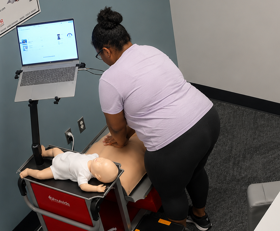 Participant performing chest compressions on an adult CPR manikin at a Citywide CPR training station in Montgomery, Alabama, with an infant CPR manikin wearing a white onesie and a laptop visible.
