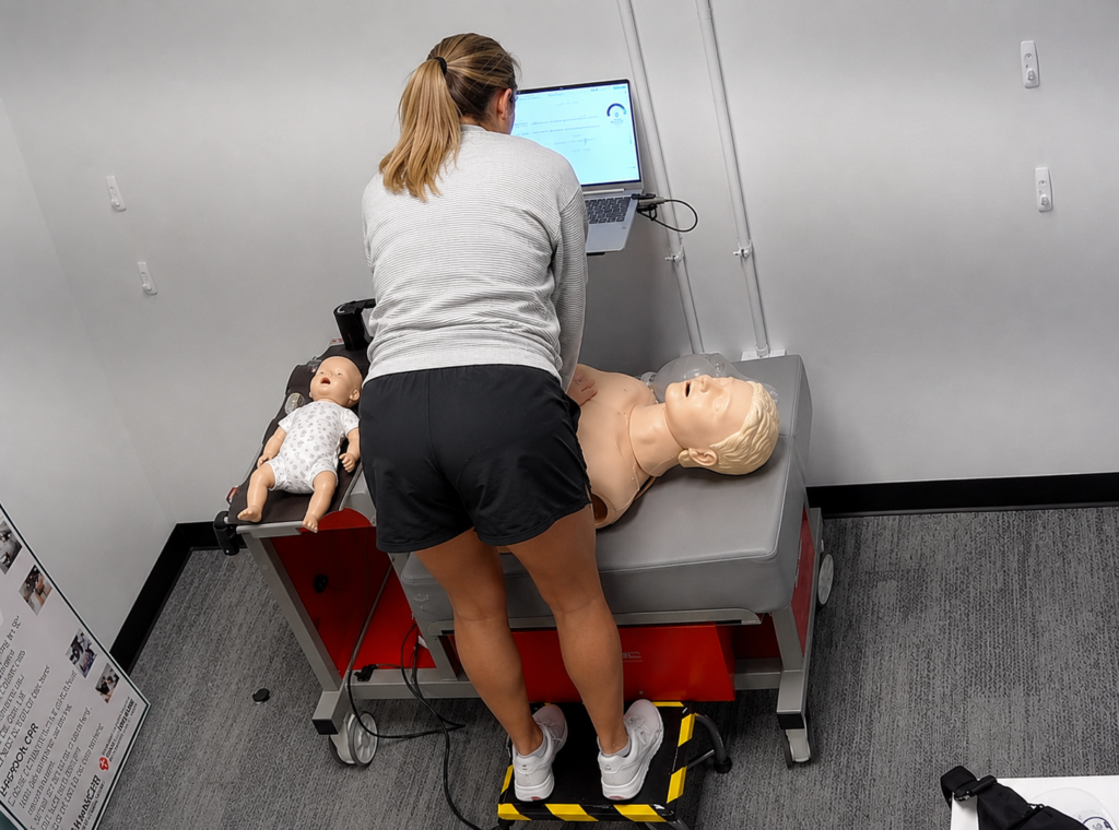 Participant performing chest compressions on an adult CPR manikin at a Citywide CPR training station in Nashville, Tennessee, with an infant CPR manikin wearing a white onesie and a laptop visible.
