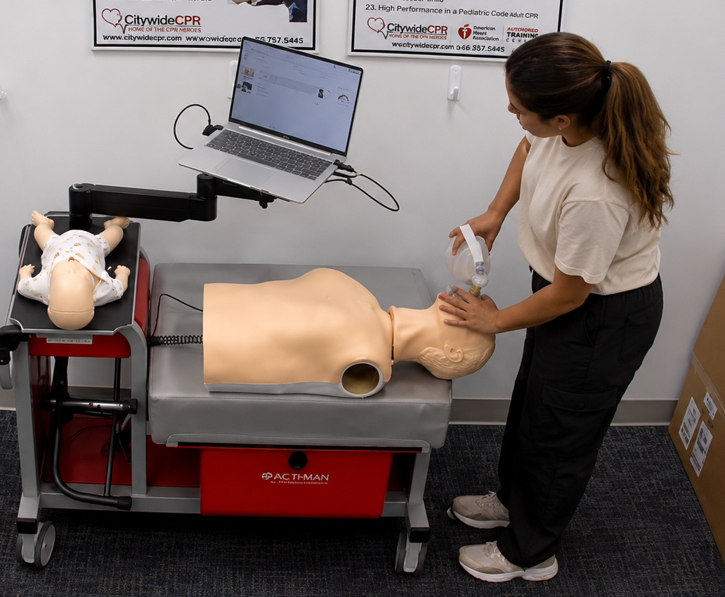 Participant providing bag-valve-mask ventilation to an adult CPR manikin at a Citywide CPR training station in Newark, New Jersey, with an infant CPR manikin wearing a white onesie and a laptop visible.
