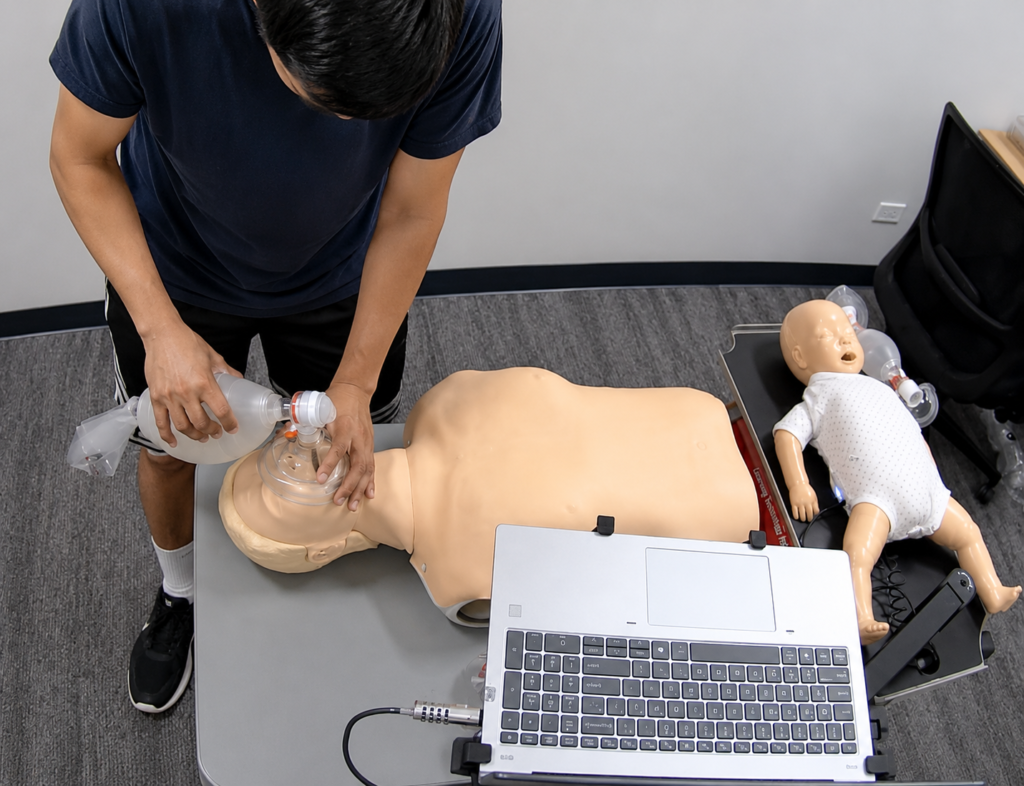 Participant providing bag-valve-mask ventilation to an adult CPR manikin at a Citywide CPR training station in Oakbrook Terrace, Illinois, with an infant CPR manikin wearing a white onesie and a laptop visible.