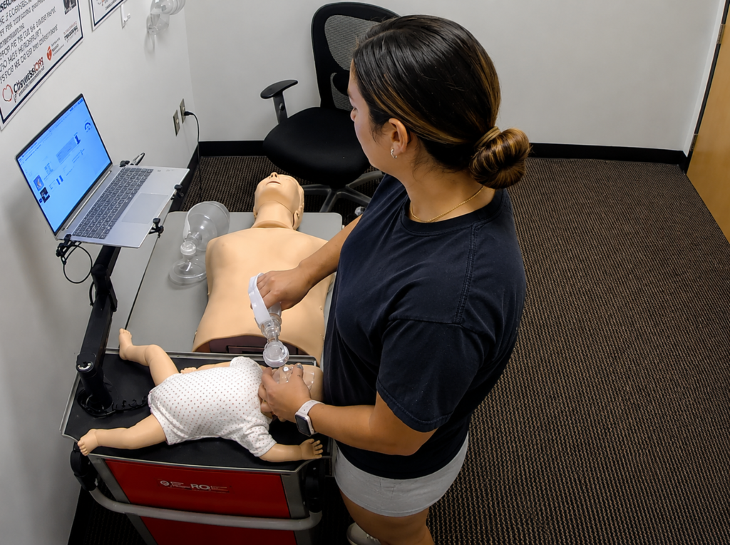 Participant providing bag-valve-mask ventilation to an infant CPR manikin wearing a white onesie at a Citywide CPR training station in Omaha, Nebraska, with an adult CPR manikin and laptop visible.