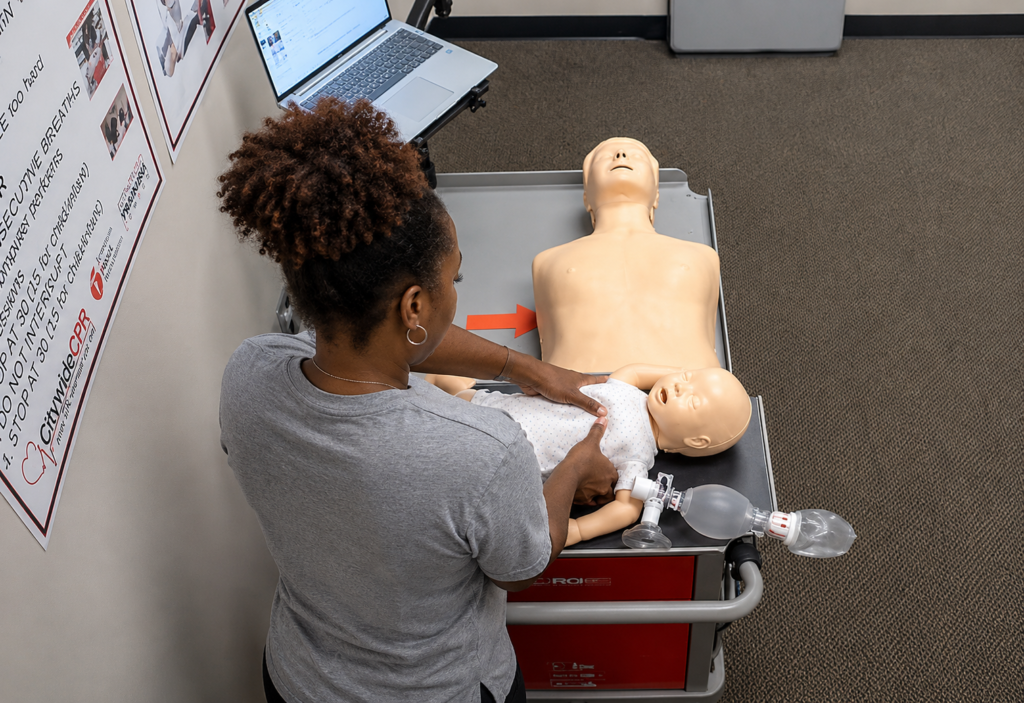 Participant performing infant CPR skills on an infant CPR manikin wearing a white onesie at a Citywide CPR training station in Orland Park, Illinois, with an adult CPR manikin and laptop visible.
