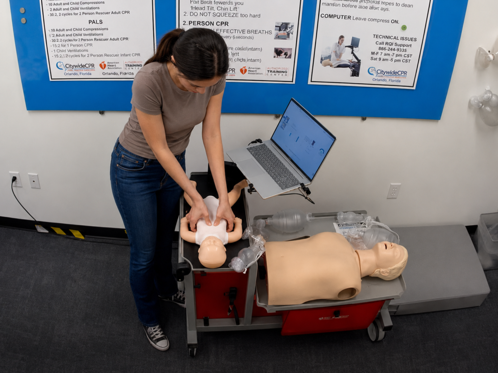 Participant performing chest compressions on an infant CPR manikin wearing a white onesie at a Citywide CPR training station in Orlando, Florida, with an adult CPR manikin and laptop visible.
