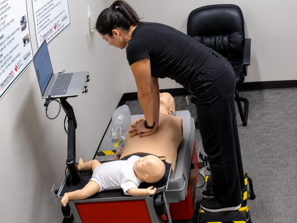 Participant performing chest compressions on an adult CPR manikin at a training station with an infant CPR manikin wearing a white onesie and a laptop nearby.