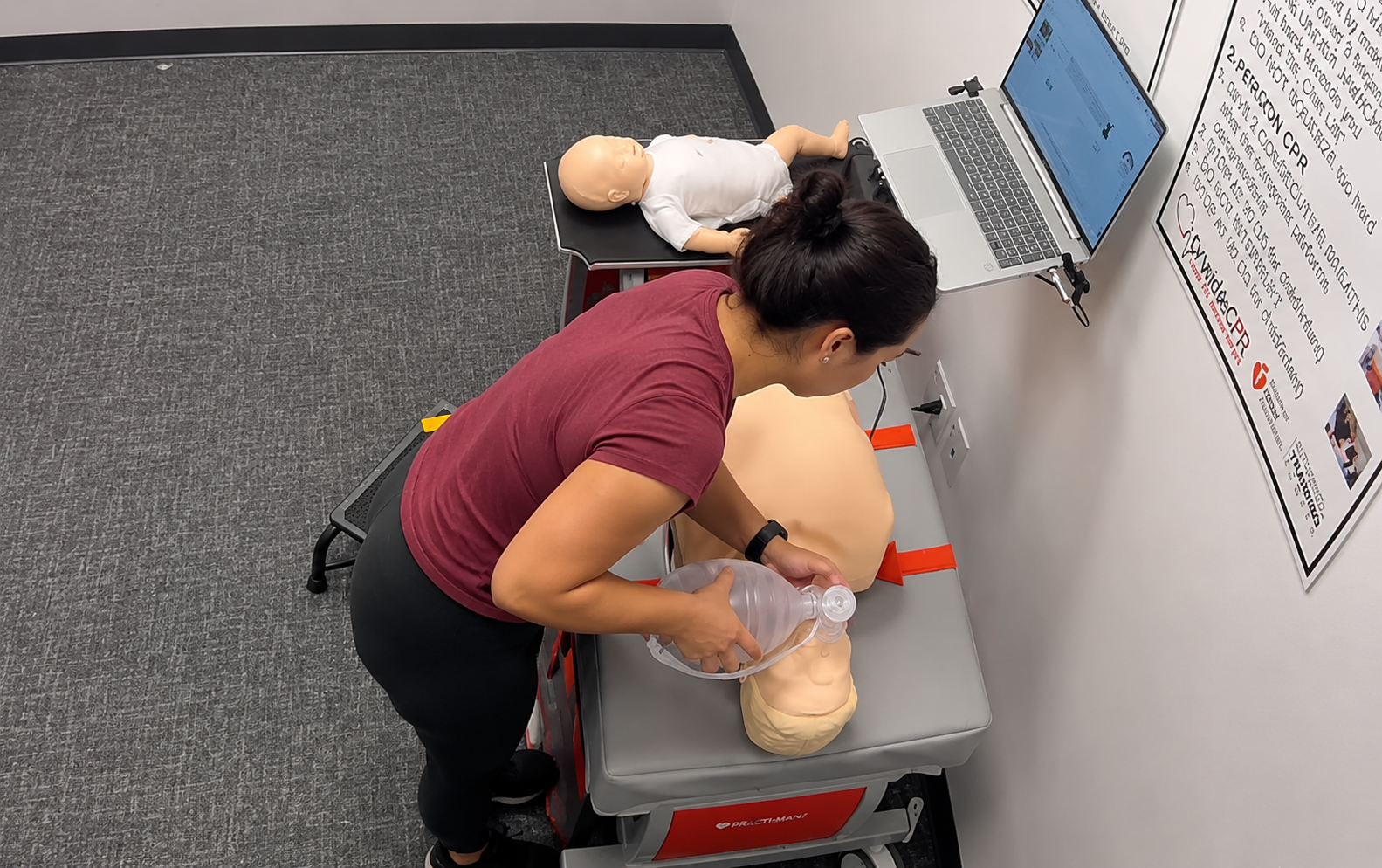 Participant providing bag-valve-mask ventilation to an adult CPR manikin at a Citywide CPR training station in San Diego, California, with an infant CPR manikin wearing a white onesie and a laptop visible.