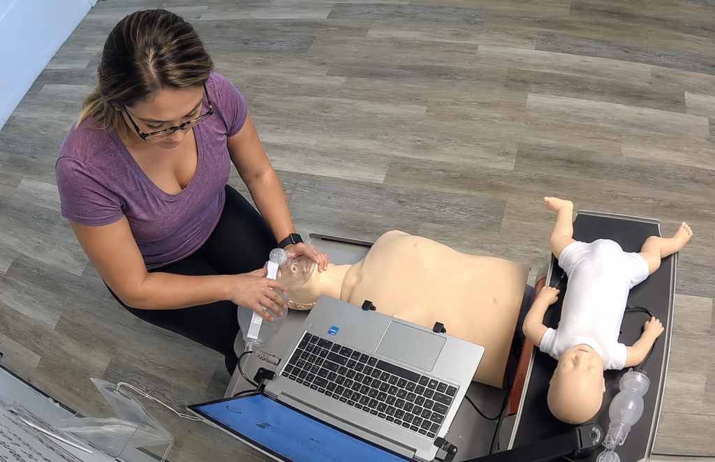Participant providing bag-valve-mask ventilation to an adult CPR manikin at a Citywide CPR training station with a laptop and an infant CPR manikin wearing a white onesie.