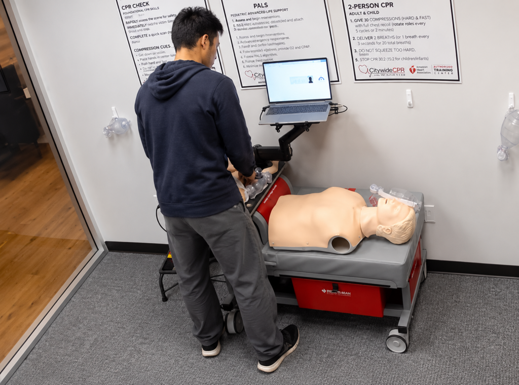 Participant standing at a Citywide CPR training station practicing airway skills on an adult CPR manikin with a laptop, CPR training posters, and ventilation equipment visible.