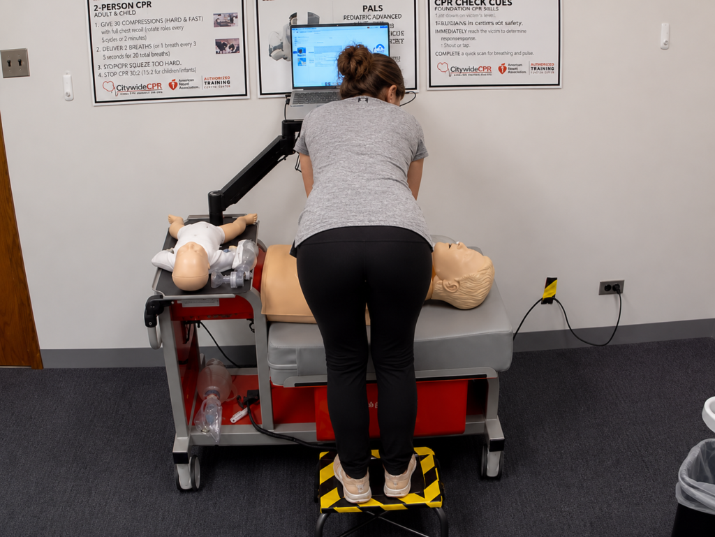 Participant performing chest compressions on an adult CPR manikin while standing on a step platform at a Citywide CPR training station, with an infant CPR manikin wearing a white onesie, a laptop, and CPR training posters visible.