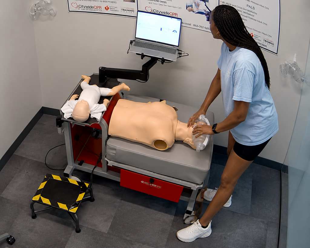 Participant providing bag-valve-mask ventilation to an adult CPR manikin at a computer-guided training station with an infant CPR manikin wearing a white onesie, a laptop, and a step platform nearby.