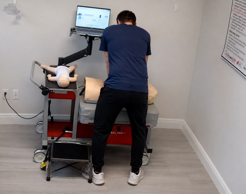 Participant performing chest compressions on an adult CPR manikin at a computer-guided training station with an infant CPR manikin wearing a white onesie, a laptop, and a step stool nearby.