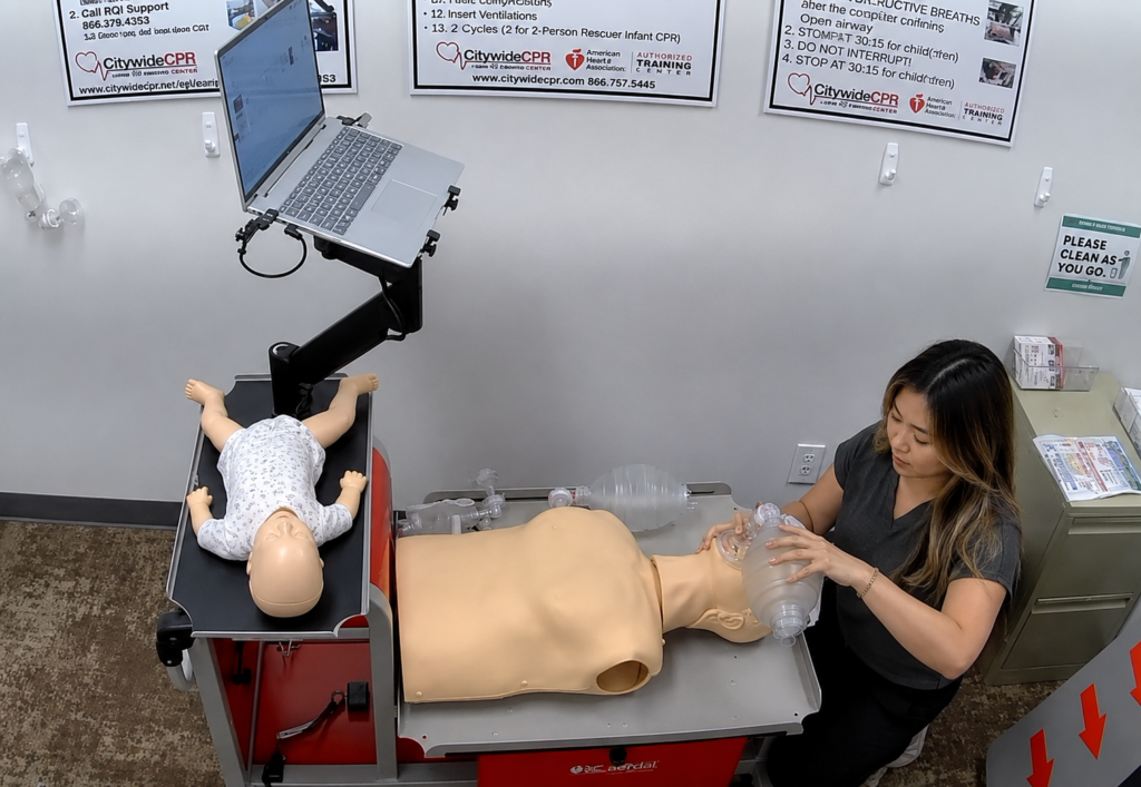 Participant providing bag-valve-mask ventilation to an adult CPR manikin at a Citywide CPR training station in Turtle Bay, New York, with an infant CPR manikin wearing a white onesie and a laptop visible.