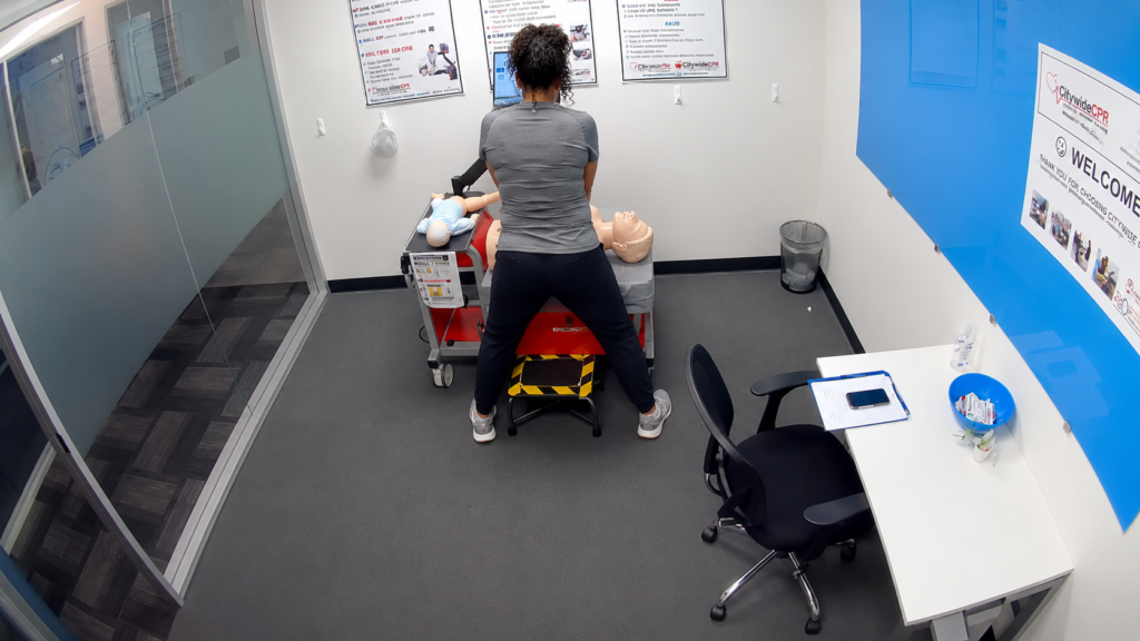 Participant standing at a Citywide CPR simulation station in West Loop, Chicago, with an adult CPR manikin, an infant manikin in a onesie, and a laptop visible.