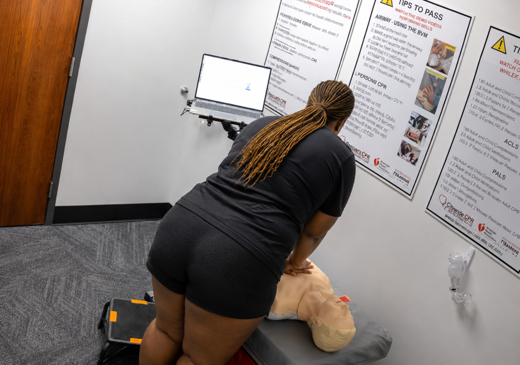 Participant performing chest compressions on an adult CPR manikin at a Citywide CPR training station in Baton Rouge, Louisiana, with a laptop visible.