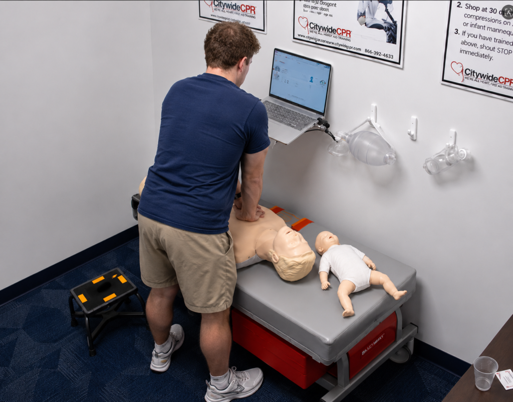 Participant performing chest compressions on an adult CPR manikin beside an infant CPR manikin wearing a white onesie, with a laptop and airway equipment visible.
