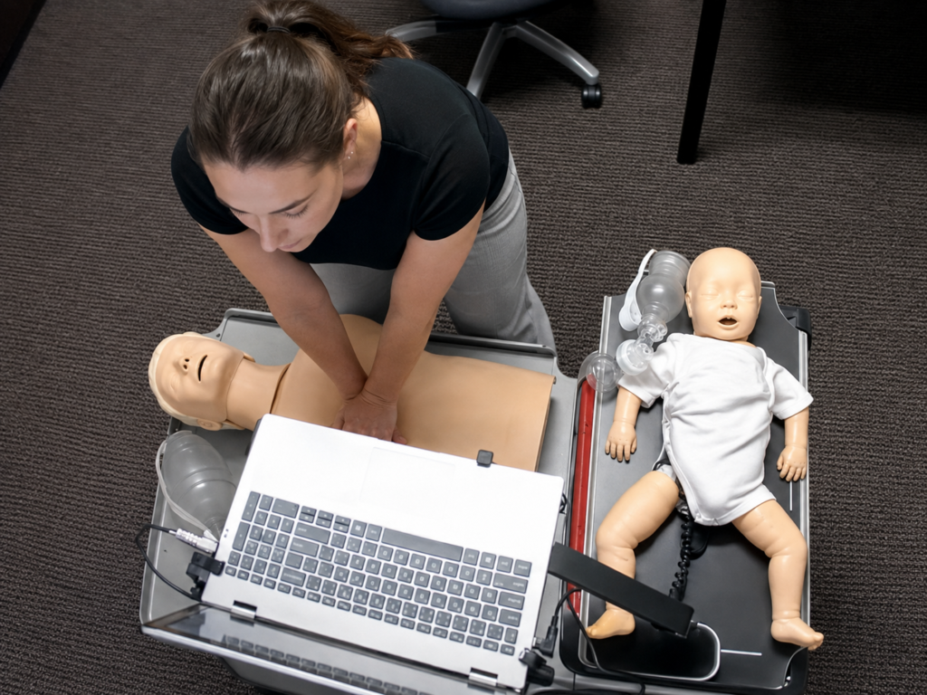 Participant performing chest compressions on an adult CPR manikin beside an infant CPR manikin wearing a white onesie, with a laptop and airway equipment visible.