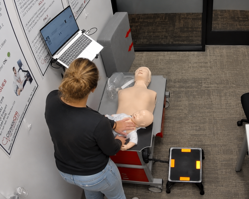 Participant performing infant CPR chest compressions on an infant CPR manikin at the Citywide CPR simulation station during American Heart Association BLS skills training in Arlington, Texas, with an adult CPR manikin and laptop visible.
