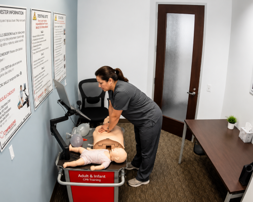 Participant performing chest compressions on an adult CPR manikin at a Citywide CPR training station in Cincinnati, Ohio, with an infant CPR manikin in a striped onesie and training equipment visible.