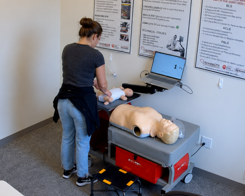 Participant performing chest compressions on an adult CPR manikin at a Citywide CPR training station in Davenport, Iowa, with an infant CPR manikin wearing a white onesie and a laptop visible.