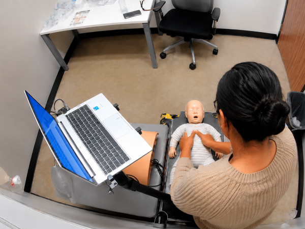 Participant performing chest compressions on a fully clothed infant CPR manikin beside a laptop at a Citywide CPR training station in Hyde Park, Chicago.