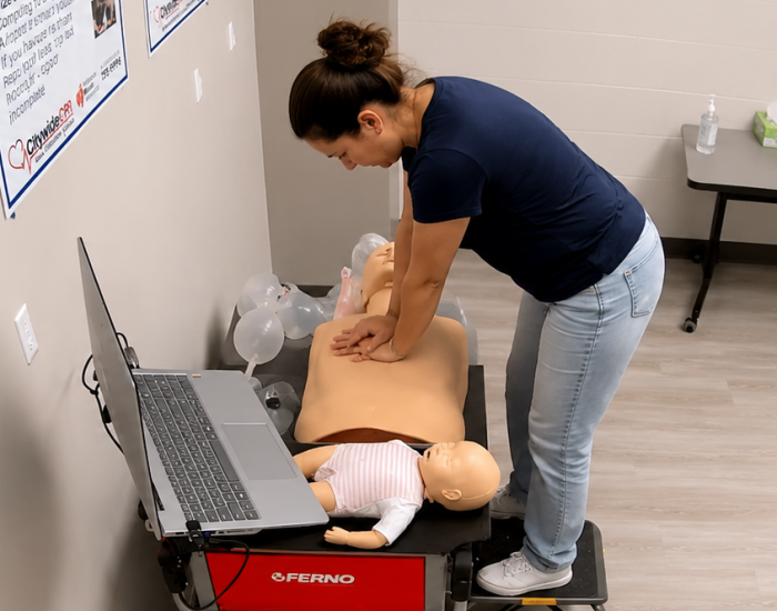 Participant performing chest compressions on an adult CPR manikin at a Citywide CPR training station in Joliet, Illinois, with an infant CPR manikin in a striped onesie and a laptop visible.