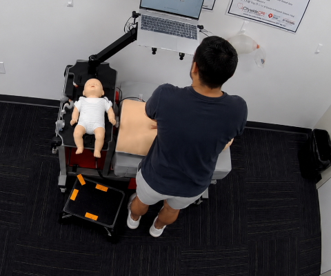 Participant using a laptop at a Citywide CPR simulation station in Boston’s Leather District, with an infant CPR manikin in a onesie and adult training manikin visible.