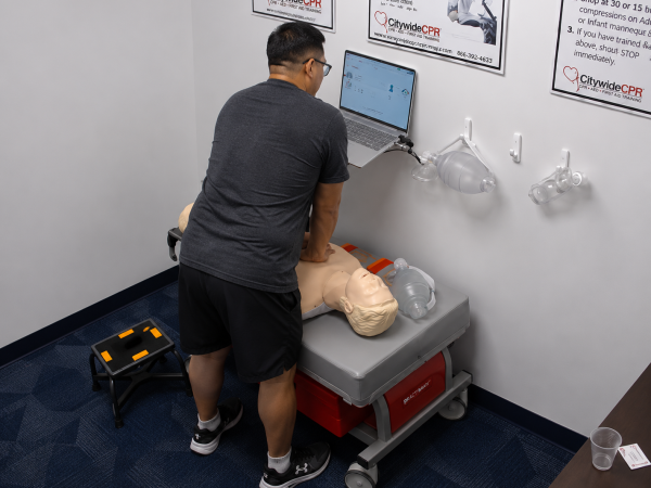 Participant performing chest compressions on an adult CPR manikin at a Citywide CPR training station in Mobile, Alabama, with a laptop and bag-valve-mask device visible.